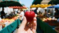 Fresh red strawberry held in hand at farmers market with produce stalls and colorful fruits in blurred background, shopping for organic food.