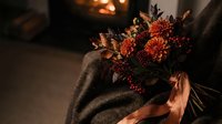 Autumn bouquet with orange dahlias, red berries, and dried leaves resting on a wool blanket by a warm glowing fireplace.