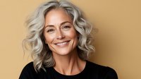 With silver gray wavy hair and natural makeup wearing black top, smiling warmly against beige background. For beauty and lifestyle content.
