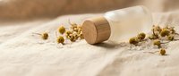 Natural chamomile flowers spilling from glass bottle with wooden cork on textured linen fabric creating spa wellness aromatherapy scene for relaxation.