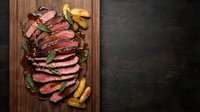 Sliced grilled beef steak on wooden cutting board with herbs and roasted vegetables, perfect for restaurant menus and culinary presentations.
