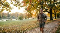 Middle-aged Caucasian man jogging through autumn park at sunrise with golden fall foliage and scattered leaves on path for fitness and wellness content.