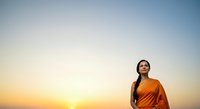 Young woman with dark hair and South Asian ethnicity wearing an orange garment, standing outdoors during golden hour sunset, looking upward with contemplative expression against clear sky.