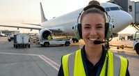 Young female airport ground crew worker with headset smiling at camera on tarmac with commercial aircraft in background during golden hour operations.