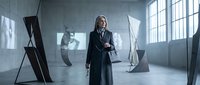 Young woman with long dark hair, wearing a dark formal blazer and tie, standing confidently in a modern industrial loft space with large windows and contemporary art installations, suitable for corporate, fashion, and lifestyle editorial use.