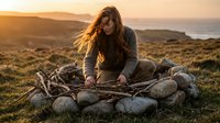 Young woman building campfire with stones at sunset in wilderness landscape with mountains and ocean view for outdoor adventure and survival camping.