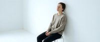 Young Asian woman in beige sweater sitting thoughtfully on white chair against minimalist background for lifestyle and wellness content.