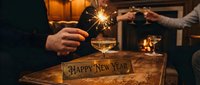 Hands holding sparklers and champagne glasses near a Happy New Year sign on a wooden table, celebrating with warm fireplace glow in background.