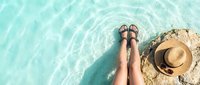 Woman legs in sandals relaxing by crystal clear turquoise water with straw hat on rocky shore. Summer vacation tropical paradise getaway concept.