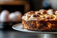Freshly baked marble cake with cream cheese swirls and walnuts on white plate, close-up view with soft natural lighting and blurred eggs in background.