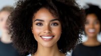 Young African American businesswoman with curly afro hair smiling confidently in modern office environment with diverse team colleagues in background.