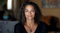 Confident mature woman with natural gray curly hair and bright smile wearing black sweater and silver pendant necklace against blurred home interior background.