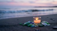 Glowing candle surrounded by sea glass on sandy beach at sunset with soft waves and pastel sky creating peaceful meditation atmosphere for wellness content.