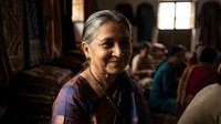 Elderly Asian woman with gray hair smiling warmly in traditional colorful sari inside ornate temple or cultural building with soft natural lighting.