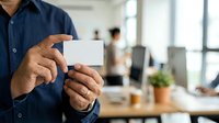Man in navy shirt holding blank white business card in modern office environment with colleagues and windows in background.