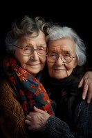 Elderly twin sisters with gray hair and glasses sharing tender moment, wearing cozy sweaters and scarves. Close-up portrait shows genuine smiles and warm embrace.