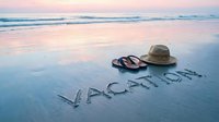 Vacation written in wet sand at sunset beach with straw hat and flip flops, pastel sky reflecting on shore for travel and holiday concepts.