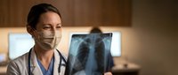 Female doctor in surgical mask examining chest x-ray film in hospital setting. Medical professional analyzing lung radiograph for diagnosis and treatment planning.