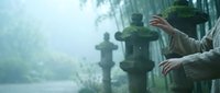 Person in gray hoodie sitting on park bench in misty morning atmosphere with blurred figures and lamp posts creating peaceful urban solitude scene.