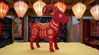 Red paper cut goat zodiac symbol with golden ornate patterns standing on wooden surface with colorful Asian lanterns hanging in traditional interior background.