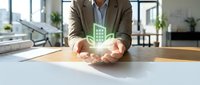 Businessman in brown blazer holding glowing green building hologram above desk in modern office with windows and plants, representing real estate development and sustainable architecture.