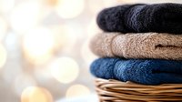 Neatly folded soft cotton towels in navy blue, beige and black stacked on wicker basket against blurred warm bokeh background, fresh laundry concept.