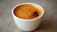 Fresh espresso with golden crema and cocoa powder in white ceramic cup on rustic surface, shallow depth of field creates soft background blur.