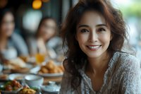 Young Asian woman with radiant smile dining at restaurant, wearing white lace top. Natural lighting creates warm ambiance with blurred background of friends gathering.