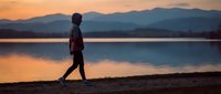 Young woman walking alone along a lake shore at sunset, silhouette against golden sky and mountain reflections on calm water.