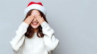 Young woman in Santa hat covering eyes with hands, wearing white sweater against gray background. Playful holiday portrait expressing joy and surprise.