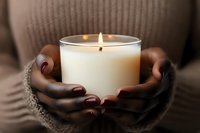 African American woman's hands with burgundy manicure holding glowing white scented candle in glass jar, creating cozy atmospheric mood for relaxation and wellness.
