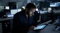 Focused businessman working late night in modern office using tablet computer analyzing data in dimly lit workspace with multiple monitors background.