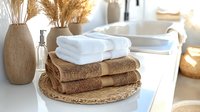 Folded brown and white cotton towels stacked on woven placemat with ceramic vase and dried pampas grass, bright sunlit bathroom interior setting.