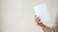 Person holding blank white paper sheet in hand against neutral beige background, suitable for mockups, presentations, and advertising campaigns.
