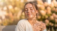 Young Caucasian woman with closed eyes enjoying warm sunlight through tree shadows on her face, peaceful moment outdoors in golden hour light for wellness content.
