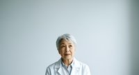 Senior Asian female healthcare professional wearing white lab coat against neutral background, suitable for medical, wellness, and healthcare industry applications.