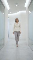 Mature woman with gray hair wearing cream blazer and taupe pants walking through modern white corridor with contemporary lighting and glass partitions, corporate environment.