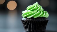 Dark chocolate cupcake with bright green buttercream frosting swirled on top, decorated with silver sprinkles against blurred bokeh background.
