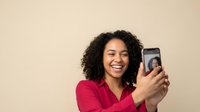 Happy young African American woman with curly hair taking selfie with smartphone against beige background for social media and communication content.