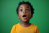 Young African child wearing yellow t-shirt expressing surprise and wonder with mouth open and eyes looking up against green background, capturing pure emotion and curiosity.