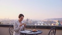 Young professional woman enjoying morning coffee on rooftop terrace overlooking historic European city skyline at sunrise with pastries and documents.