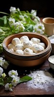 Steamed dumplings in bamboo basket with white flowers and tea cup on wooden table. Dark moody food photography with traditional Asian cuisine setting.