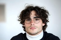 Young Hispanic man with curly dark hair and round glasses wearing white turtleneck sweater looking confidently at camera against light background.
