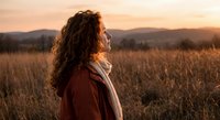 Young woman with curly hair enjoying peaceful sunset moment in golden field, looking up with gratitude and serenity during golden hour outdoor meditation.