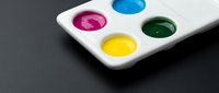 Watercolor palette with vibrant magenta, blue, yellow, and green paint wells on white ceramic tray against dark background for art and design projects.