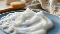Creamy white foam cleanser or soap bubbles on blue plate with natural bath accessories creating luxurious spa skincare routine for relaxation and wellness.