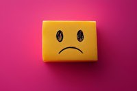 Yellow sponge with sad face drawn on it against vibrant pink background, representing depression, sadness, and negative emotions in minimalist style.