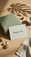 Thank you card leaning against sage green gift box on wooden surface with dried flowers and monstera leaf shadow in warm natural light.
