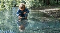 Young Caucasian boy crouching by a shallow stream, touching water with curiosity, surrounded by lush green trees in summer nature.