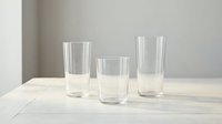 Three empty clear glass tumblers of varying heights arranged on light wooden surface against neutral gray wall background for kitchen decor.